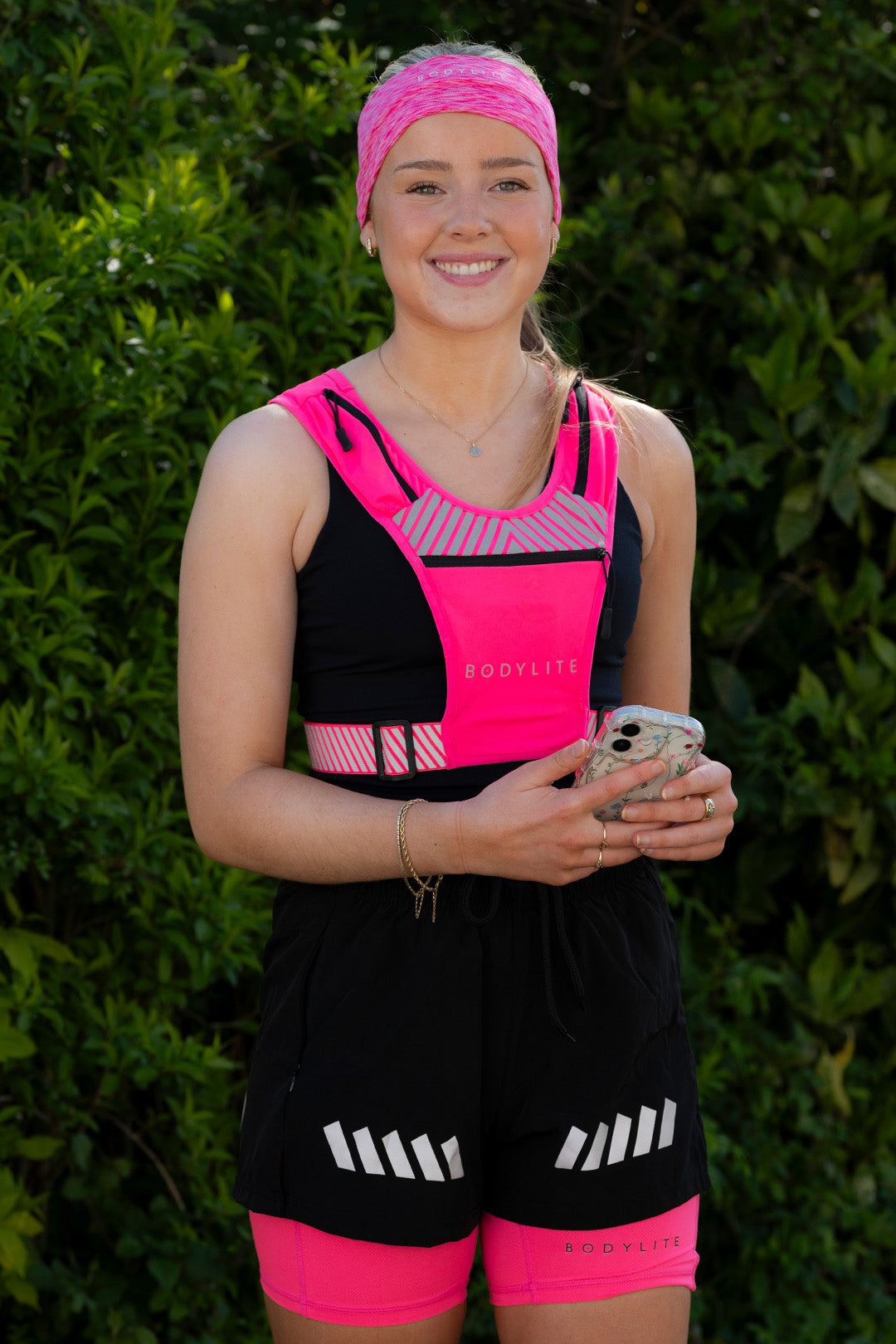 A person wearing a neon pink and black phone holder vest and shorts with reflective details. They are smiling and holding a phone.