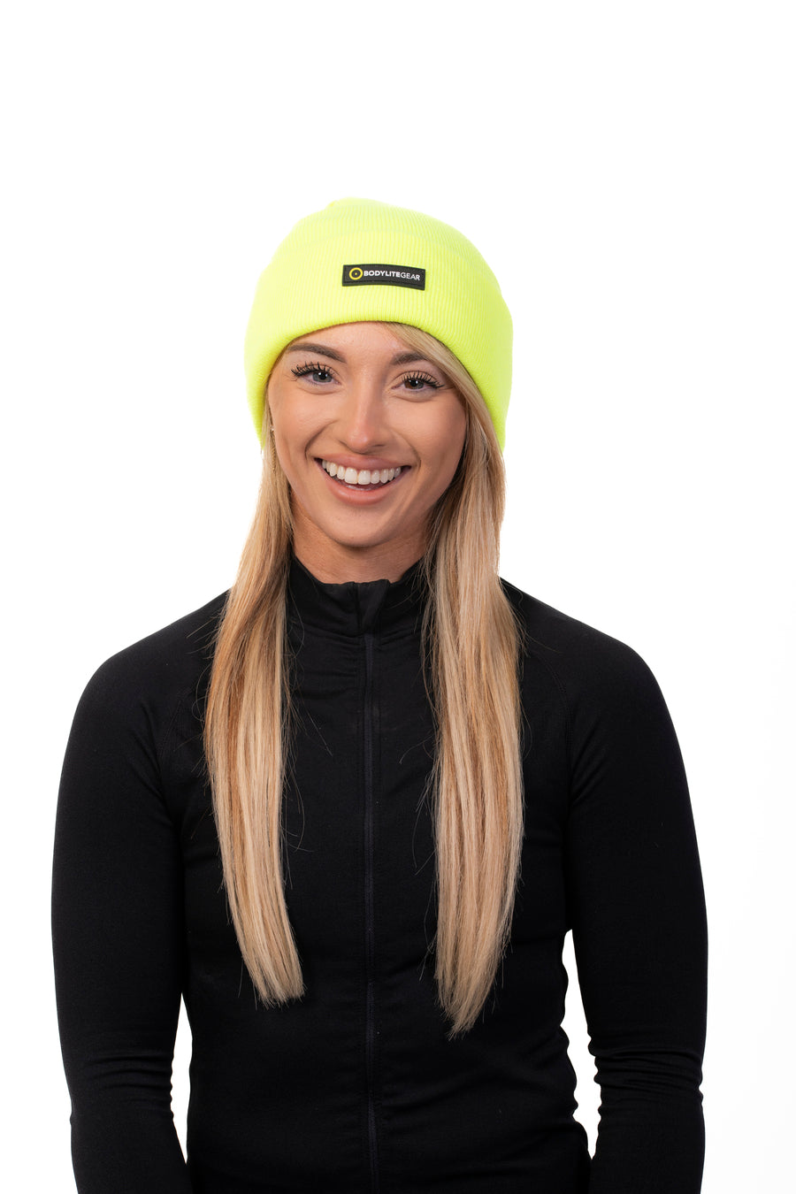 A woman wearing a neon yellow beanie and a black jacket. She is smiling and standing against a white background.