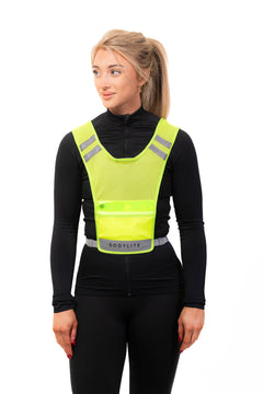 A person wearing a yellow LED reflective vest with adjustable straps and a front pocket, over a black top and pants.